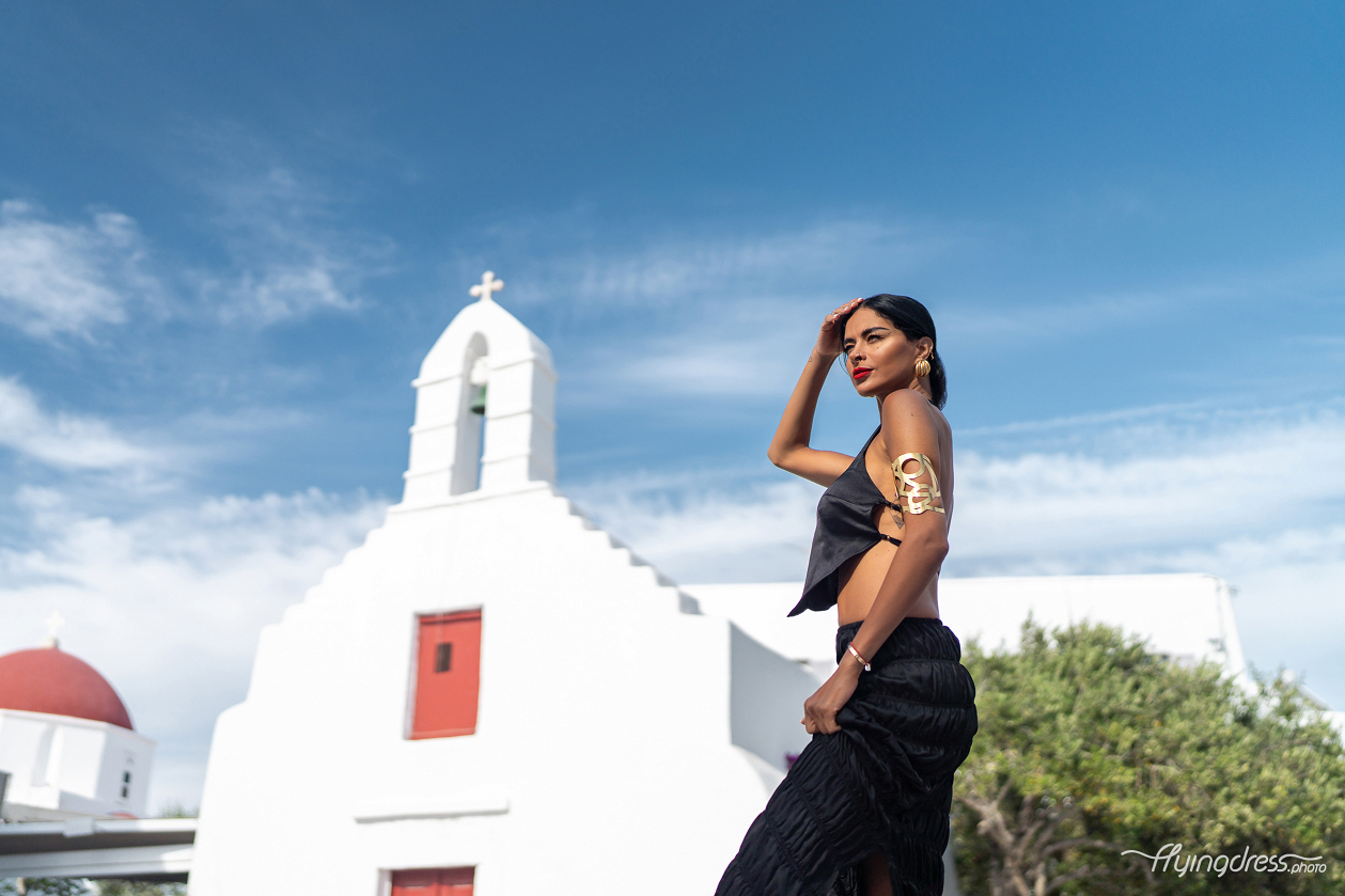 A woman in a sleek black outfit poses confidently against a backdrop of a white church with red accents in Mykonos, under a clear blue sky.